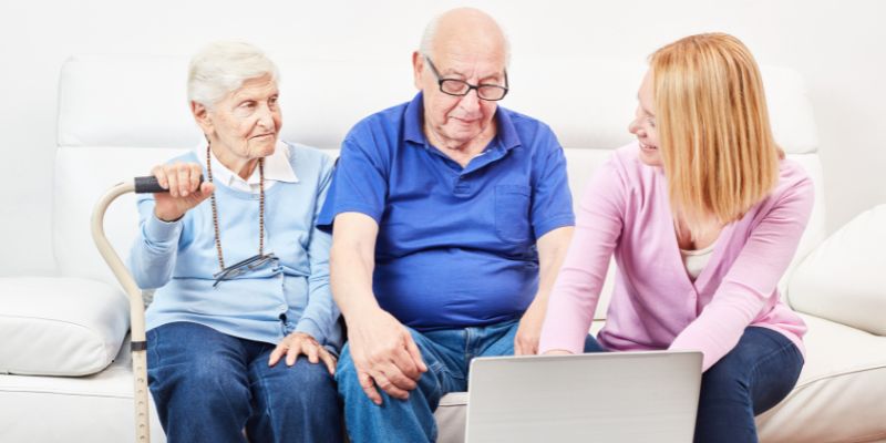 Elderly couple sitting on a couch with a younger woman showing them something on a laptop, learning how to apply for social security benefits at age 62 together as a family.