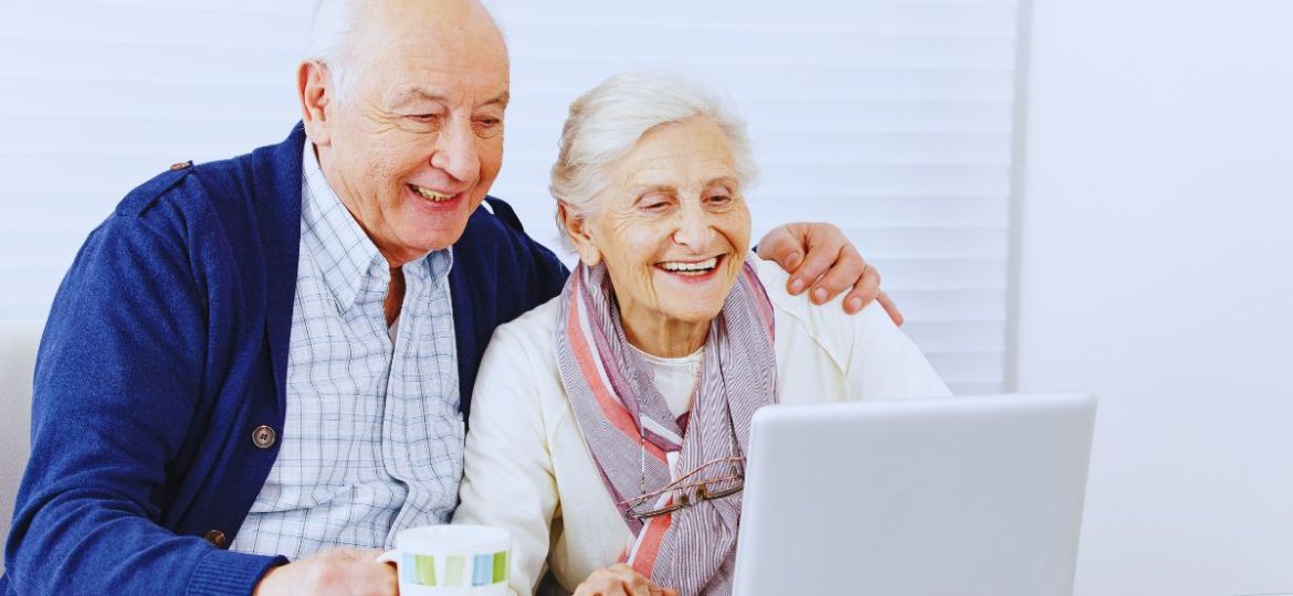 Happy senior man and woman sharing a laptop on the couch, with the man holding a coffee mug while they explore how to apply for social security benefits at age 62 online.