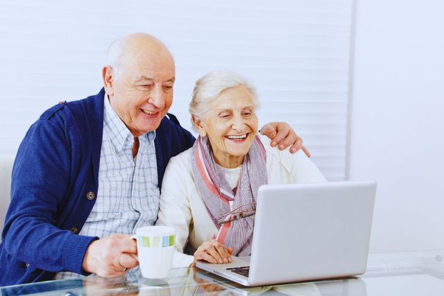 Happy senior man and woman sharing a laptop on the couch, with the man holding a coffee mug while they explore how to apply for social security benefits at age 62 online.