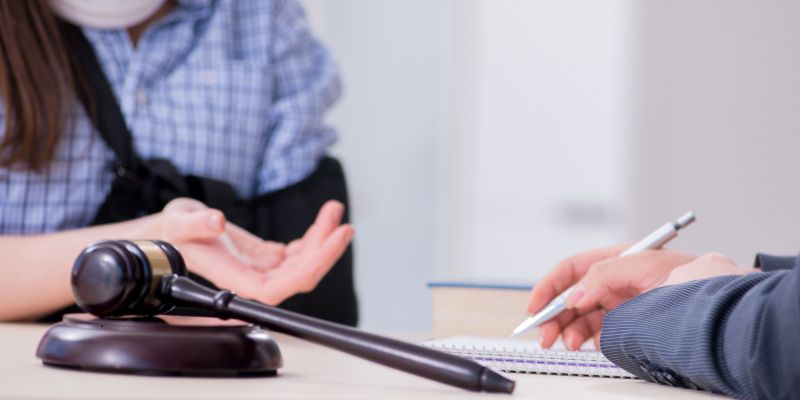 LOP1 Injured client wearing an arm sling and casual checkered shirt seated at a lawyer's desk, gesturing with an open palm while the attorney takes notes, with a wooden judge's gavel prominently displayed in the foreground, representing the initial consultation where a LOP (Letter of Protection) is often discussed to secure medical treatment in a personal injury case