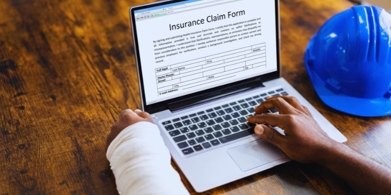does-workers-comp-pay-for-lost-wages2 Worker with bandaged arm filling out an online insurance claim form on a laptop, with safety helmet beside, illustrating the process when wondering does workers comp pay for lost wages after a workplace injury